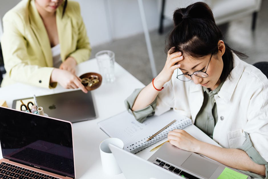 A woman in glasses stressed over work at a laptop, holding her head in an office setting.